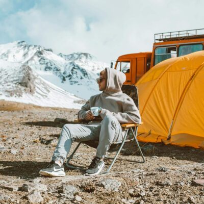 Full length of male camper sitting on folding chair next to tent in middle of mountain valley and enjoying views