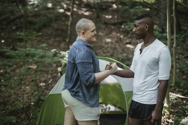 Positive bald guy giving hand shake to black friend while standing in forest near tent on sunny day