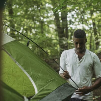 Serious young African American guy in white shirt inserting stick of tent in hole during hiking in summer forest