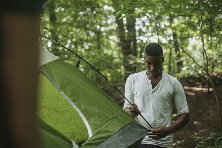 Serious young African American guy in white shirt inserting stick of tent in hole during hiking in summer forest