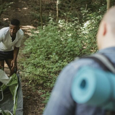 Unrecognizable male backpacker standing and looking at African American guy in shorts setting up tent in forest on sunny day